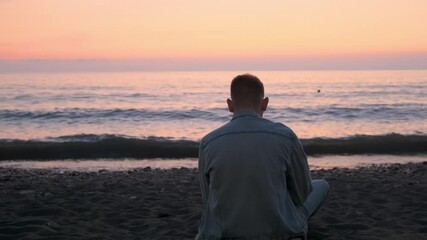 Rear view of Caucasian man sitting alone on black sand beach on the coast and enjoying beautiful sunset over the sea. Thoughtful male traveler enjoy freedom and tranquility of contemplating nature