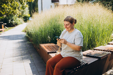 Fototapeta premium Happy woman chatting on phone and relaxing on the bench outdoors. Urban people concept. Smiling woman text messaging on smart phone at sunny day.