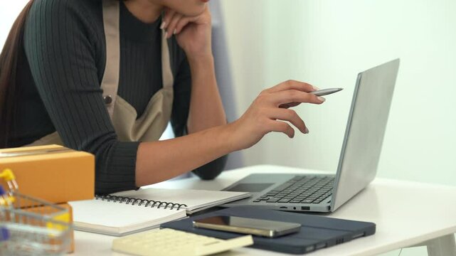 Focused Online Business Owner: A female entrepreneur thoughtfully works on her laptop, surrounded by packaging materials and small business tools.  The image portrays dedication and concentration. 