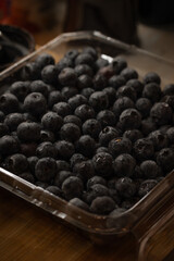 a plastic container filled with fresh blueberries. The container is likely sitting on a wooden surface, given the visible wood grain in the background