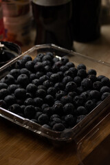 a plastic container filled with fresh blueberries. The container is likely sitting on a wooden surface, given the visible wood grain in the background
