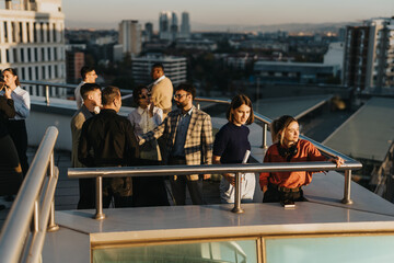 A diverse group of business colleagues brainstorm project solutions while standing on a high tower balcony, demonstrating teamwork and innovative thinking during a sunset meeting in an urban setting.