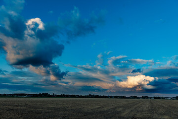 Dramatic cloudscape over a plowed field, with a small town in the distance.  Evening light illuminates puffy clouds against a deep blue sky.