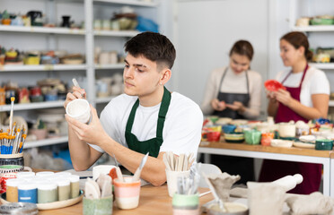 Young man painting clay ceramic cup in workshop