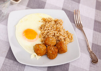 Hearty breakfast setup showing creamy oatmeal with sunny side up egg and savory meatballs on white plate, arranged on checkered tablecloth
