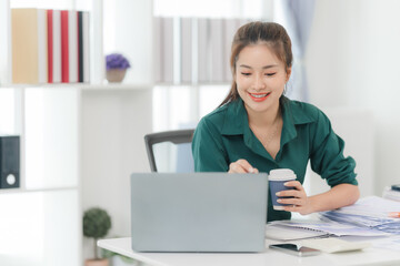 Focused and Productive: Young businesswoman working efficiently at her desk, laptop and coffee in hand.  A picture of modern workplace success. 