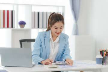 Focused Businesswoman Analyzing Data: A portrait of a young, successful Asian businesswoman meticulously reviewing financial documents at her desk, showcasing professionalism and dedication.