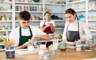 Interested young man and woman in pinafores sculpting handmade jars of potter-earth material in art studio