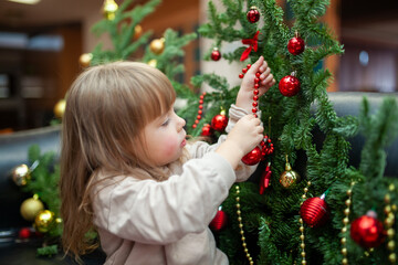 A young child carefully decorates a Christmas tree with red ornaments and festive beads. The holiday scene captures the joy of childhood and the magic of preparing for Christmas in a cozy indoor