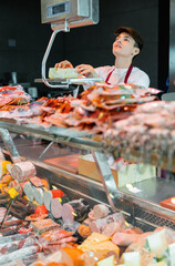 Young man in apron working in cheese and meat shop, standing at counter and using scale to weigh piece of cheese.