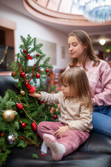 A young child decorates a small Christmas tree with bright red ornaments under the watchful eye of a caring older sibling. A cozy holiday moment highlighting family, tradition, and festive cheer.