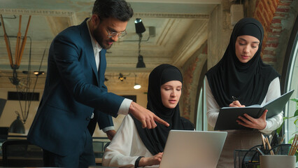 Fototapeta premium Three coworkers colleagues businessman businesswomen collaborate work together Arabian man muslim women hijab male female teamwork discuss talk write typing laptop business project at office company