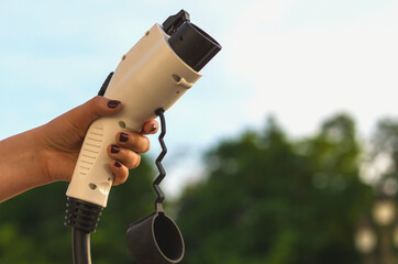 A female hand holds a charging cable of an electric car against the background of the sky and nature. The driver holds a cable for charging an electric vehicle. Clean alternative energy concept..