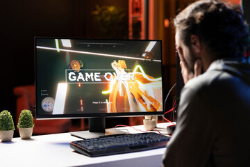 Player upset after losing while playing first person shooter videogame, putting face in hands in frustration. Man feeling dejected after receiving game over screen while playing on PC, crying