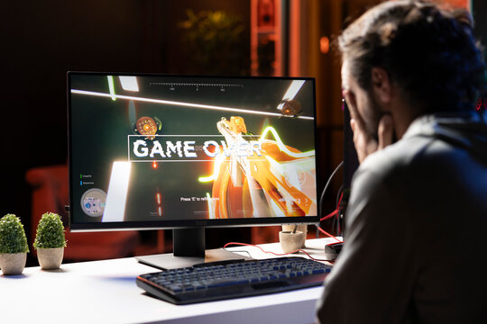 Player upset after losing while playing first person shooter videogame, putting face in hands in frustration. Man feeling dejected after receiving game over screen while playing on PC, crying