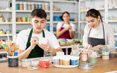 Concentrated young man in apron painting on handmade clay cup with brush in pottery workshop....