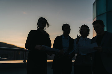 A group of business people engaged in a meeting on a rooftop, silhouetted against the setting sun, creating an atmosphere of collaboration and teamwork. Ideal for business and teamwork concepts.