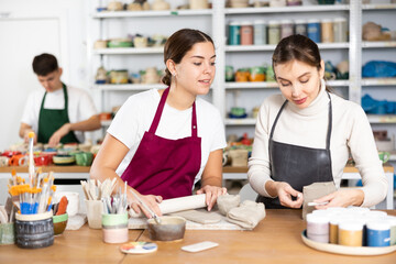 Cheerful young women in aprons modeling handmade mugs of row clay material in pottery workroom