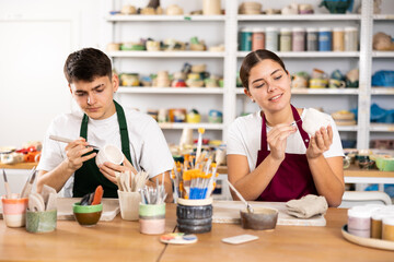 Concentrated young man and woman in aprons engraving on clay cup with cutter in pottery workshop