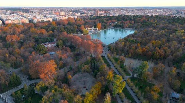 Breathtaking aerial view of El Retiro Park beautifully captures the vivid fall foliage along with the peaceful lake, all while featuring the urban sunset landscape of Madrid rising in the background