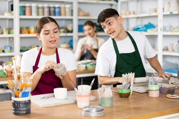 Smiling young man and woman in aprons making handmade cups of row clay material in pottery workshop