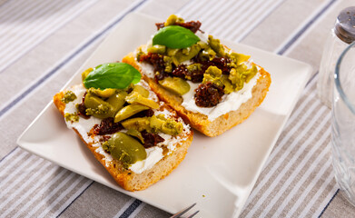 Appetizing sandwich with curd cheese, sun-dried tomatoes, pesto sauce and olives on a white square plate on the tablecloth in a cafe