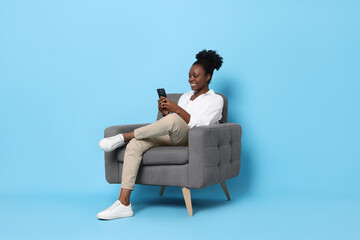 Smiling woman with smartphone sitting in armchair on light blue background