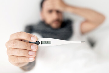 Sick young man showing thermometer to camera, lying on bed and touching forehead. Coronavirus, influenza symptom concept. Selective focus, top view