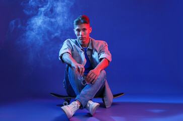 Fototapeta premium Handsome man sitting on skateboard in smoke and color lights against blue background