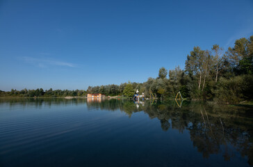 A blue lake against a blue sky. There are trees in the background. At the edge of the lake, a children's playground and a wooden hut, which are partially flooded.