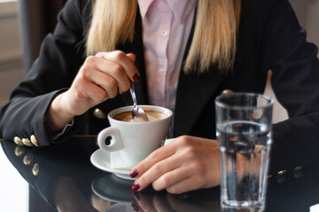 Woman hand stirring coffee with a small metal spoon on a restaurant desk. Elegant jacket visible, drink concept, close-up, selective focus