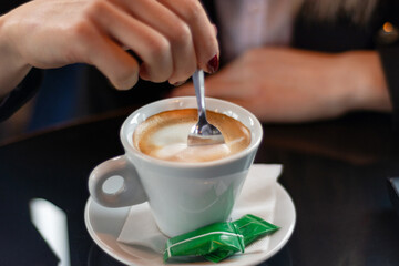 Woman's hand stirring coffee with a small metal spoon on a restaurant desk. Drink concept, close-up, selective focus