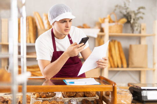Focused young bakery manager in maroon apron using smartphone and paperwork to coordinate customer orders, standing at counter in cozy shop filled with fresh bread and pastries arranged on shelves - Powered by Adobe