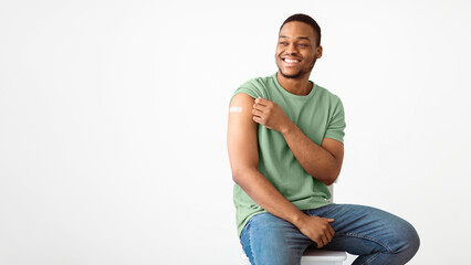 Fototapeta premium Covid-19 Vaccination. Joyful African Man Showing Vaccinated Arm With Medical Plaster After Coronavirus Vaccine Injection Sitting Over White Studio Background. Covid Immunization. Panorama, Copy Space
