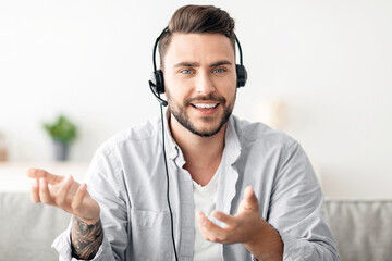 Excited man in headset talking via webcam, having video call with distant colleague working remotely, sitting on sofa at home. Video conference, modern communication, online business training concept
