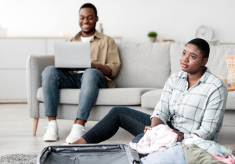 African American Wife Packing Travel Suitcase While Husband Using Laptop Browsing Internet And Buying Tickets Online Sitting At Home. Selective Focus On Black Woman Dreaming About Vacation