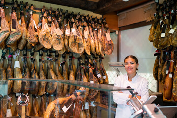 Portrait of positive female butcher on background of rack with hanging various jamon in shop