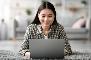 Relaxed millennial asian woman looking for job opportunities online, laying on carpet amd using modern laptop at home, smiling and typing on notebook keyboard, chinese lady working remotely, closeup