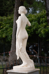 An antique statue made of white stone with a layer of old dirt stands among trees in a garden