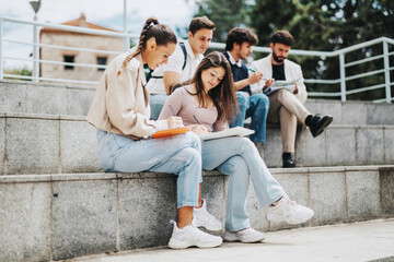 A group of university students collaborates in an outdoor setting with their professor offering guidance. They sit on steps, focusing on their notes and discussions in a supportive learning