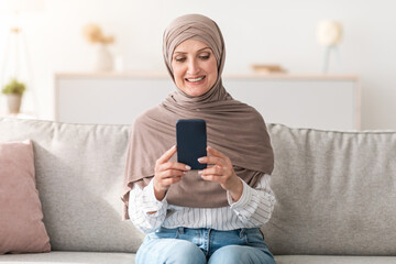 Obraz premium Happy Senior Muslim Woman Using Phone Texting And Browsing Internet Sitting On Couch At Home, Wearing Traditional Hijab. New Mobile Application For Smartphone Advertisement Concept