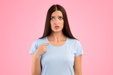 Fototapeta premium Are You Looking At Me. Portrait of confused young lady pointing finger at herself, posing at camera, standing isolated over orange studio background. Surprised scared female model indicating her