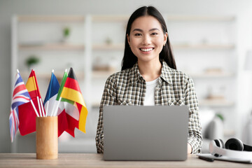 Positive asian young woman foreign language teacher sitting at workdesk with international flags...