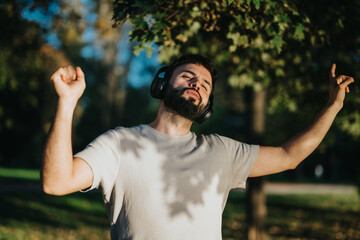 A young man wearing headphones enjoys music outdoors. He appears relaxed and content under a bright, sunny sky in a lush green park. An expression of freedom and happiness is evident.