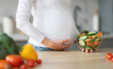 A pregnant woman stands in her kitchen, cradling her baby bump while preparing a healthy salad with fresh vegetables. Bright lights illuminate the inviting cooking space, cropped
