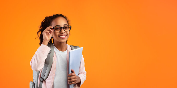 Education Concept. Portrait of positive smiling African American girl touching glasses holding textbooks, wearing backpack and looking at camera isolated on yellow studio background