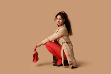 Beautiful young African-American woman in stylish red tights with handbag sitting on brown background