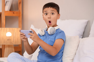 Cute shocked little boy with mobile phone and headphones playing video games at home