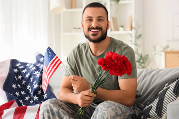 Young soldier with USA flag and carnation flowers sitting on sofa at home. Veterans Day celebration