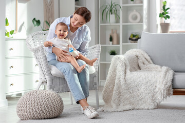 Mother giving her cute little baby bottle of milk in armchair at home